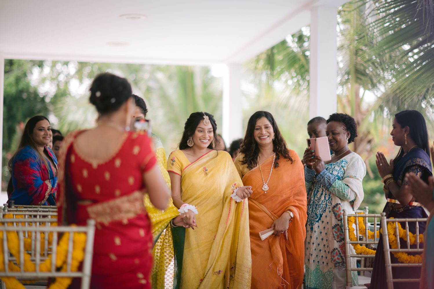 Women in colorful sarees celebrating at an outdoor Indian wedding ceremony with lush greenery in the background.