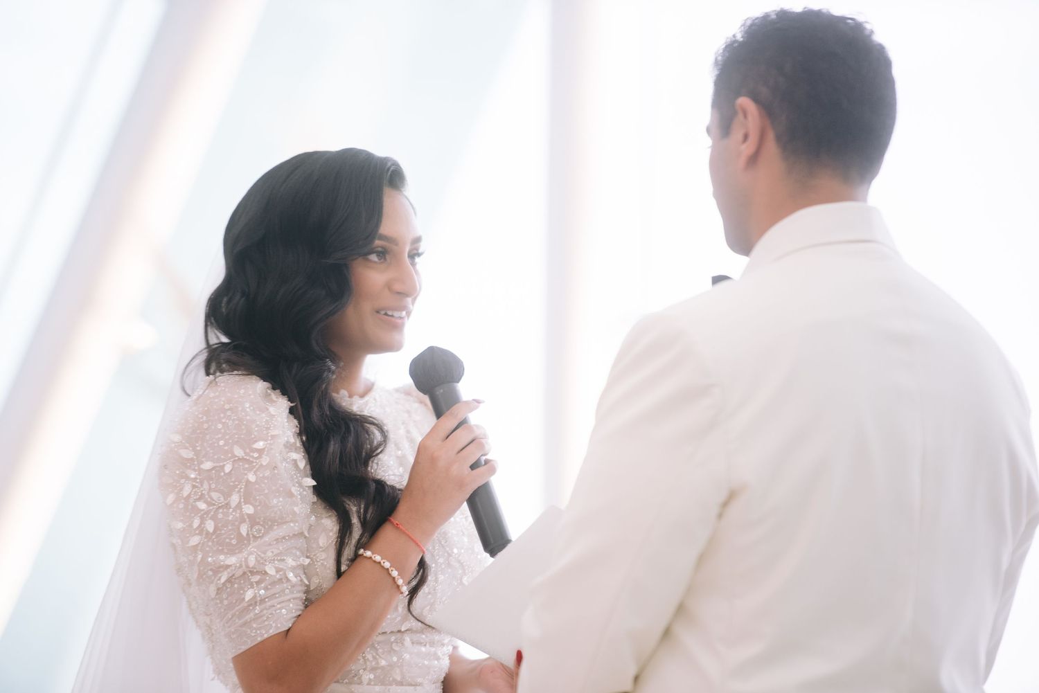 Bride holding microphone reciting wedding vows to groom in white suit during elegant ceremony.