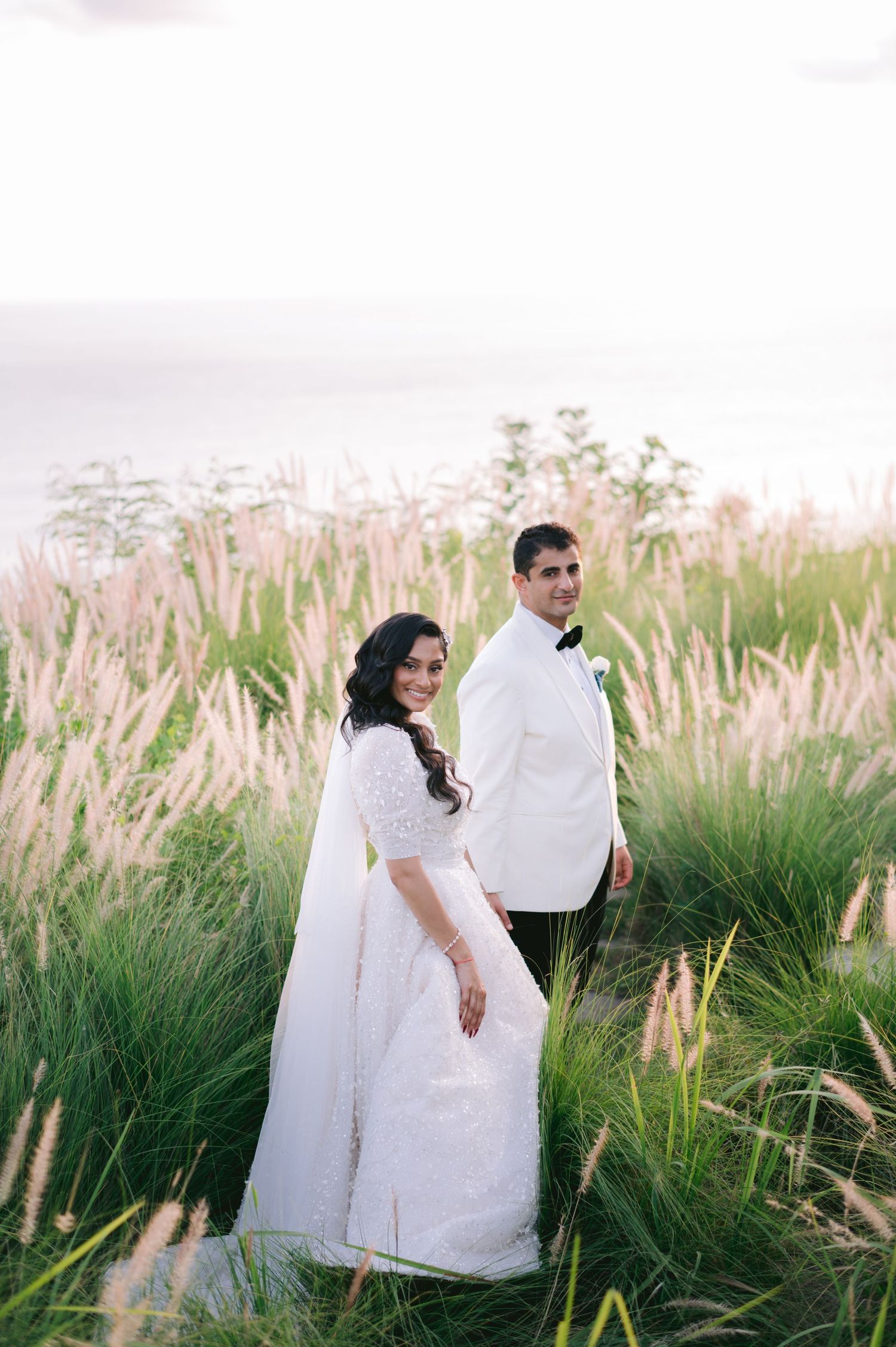 Bride in white lace gown and groom in white tuxedo standing among tall green grass at golden hour.