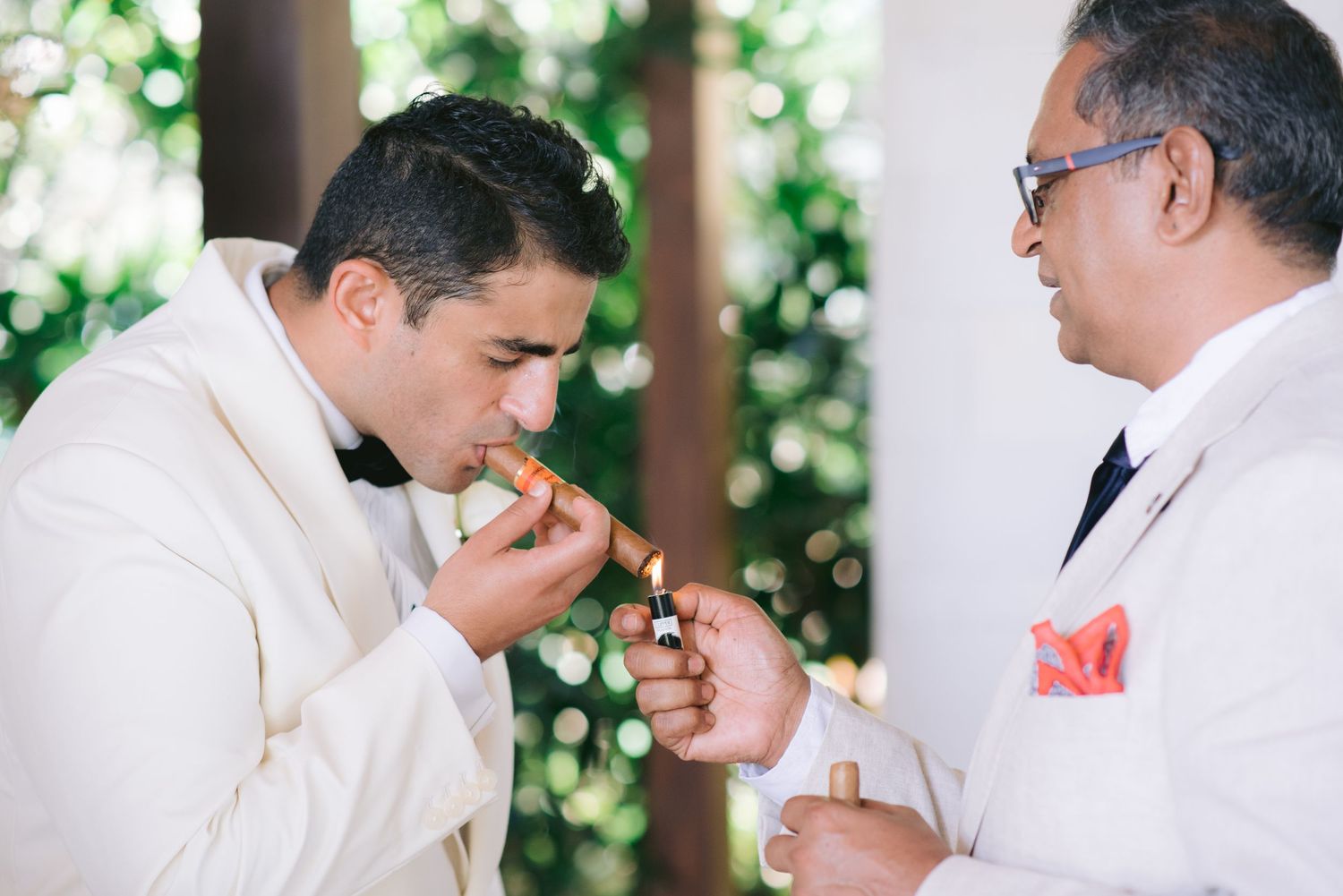 Two men in white suits lighting a cigar outdoors at a wedding celebration.