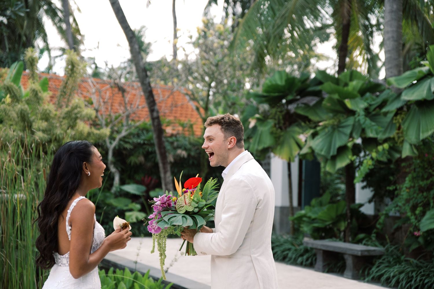 Bride and groom share a joyful first look moment outdoors, surrounded by lush tropical greenery with colorful bouquet.