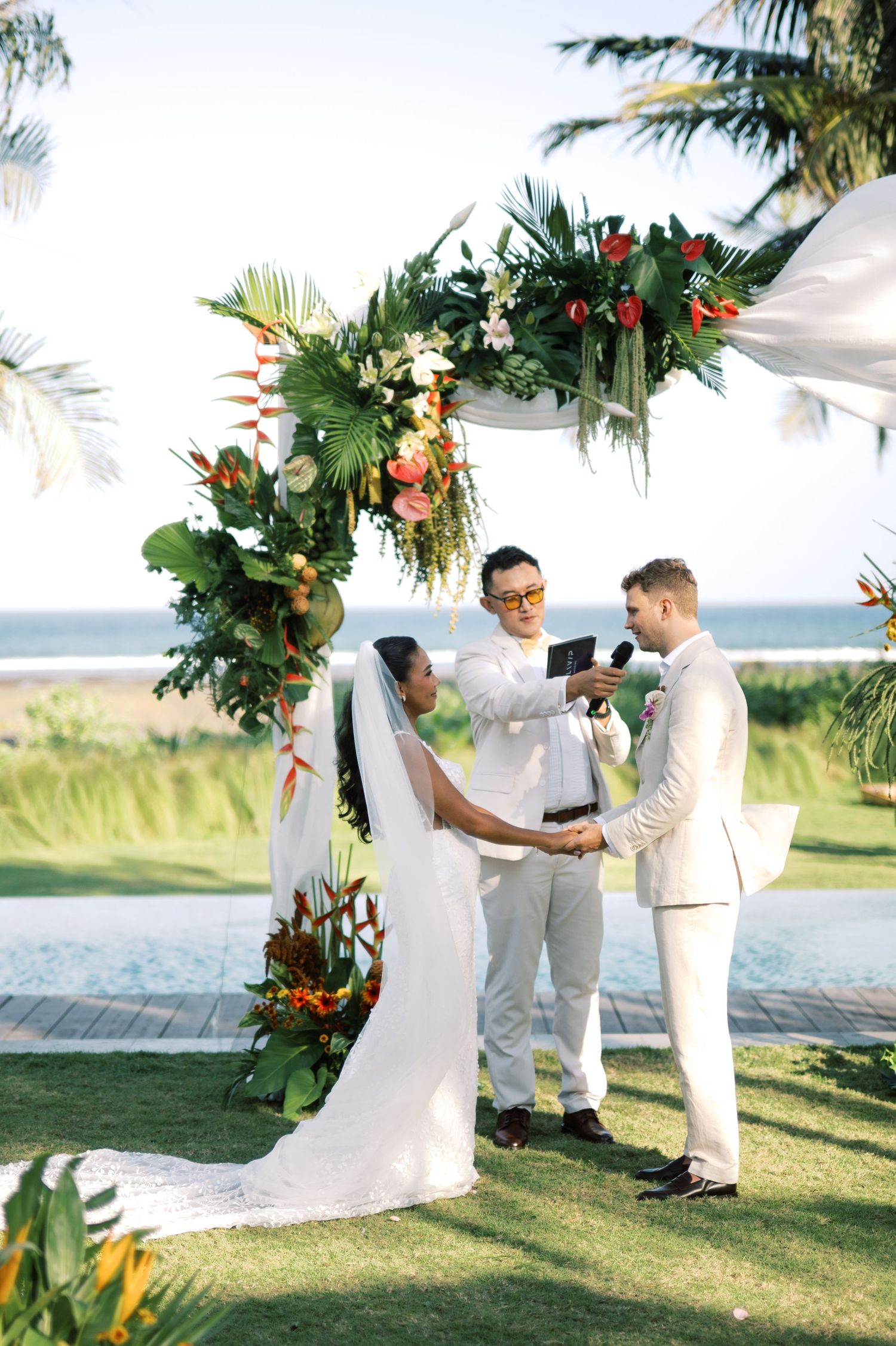 Bride and groom exchanging vows under a tropical floral arch with officiant at a beachside wedding.