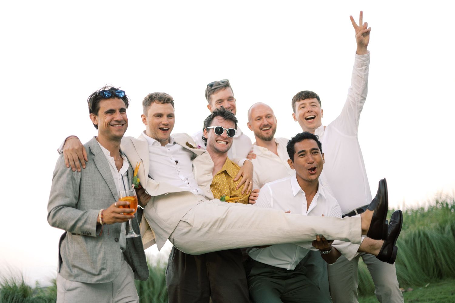 Group of groomsmen in suits laughing and lifting groom outdoors at wedding celebration.
