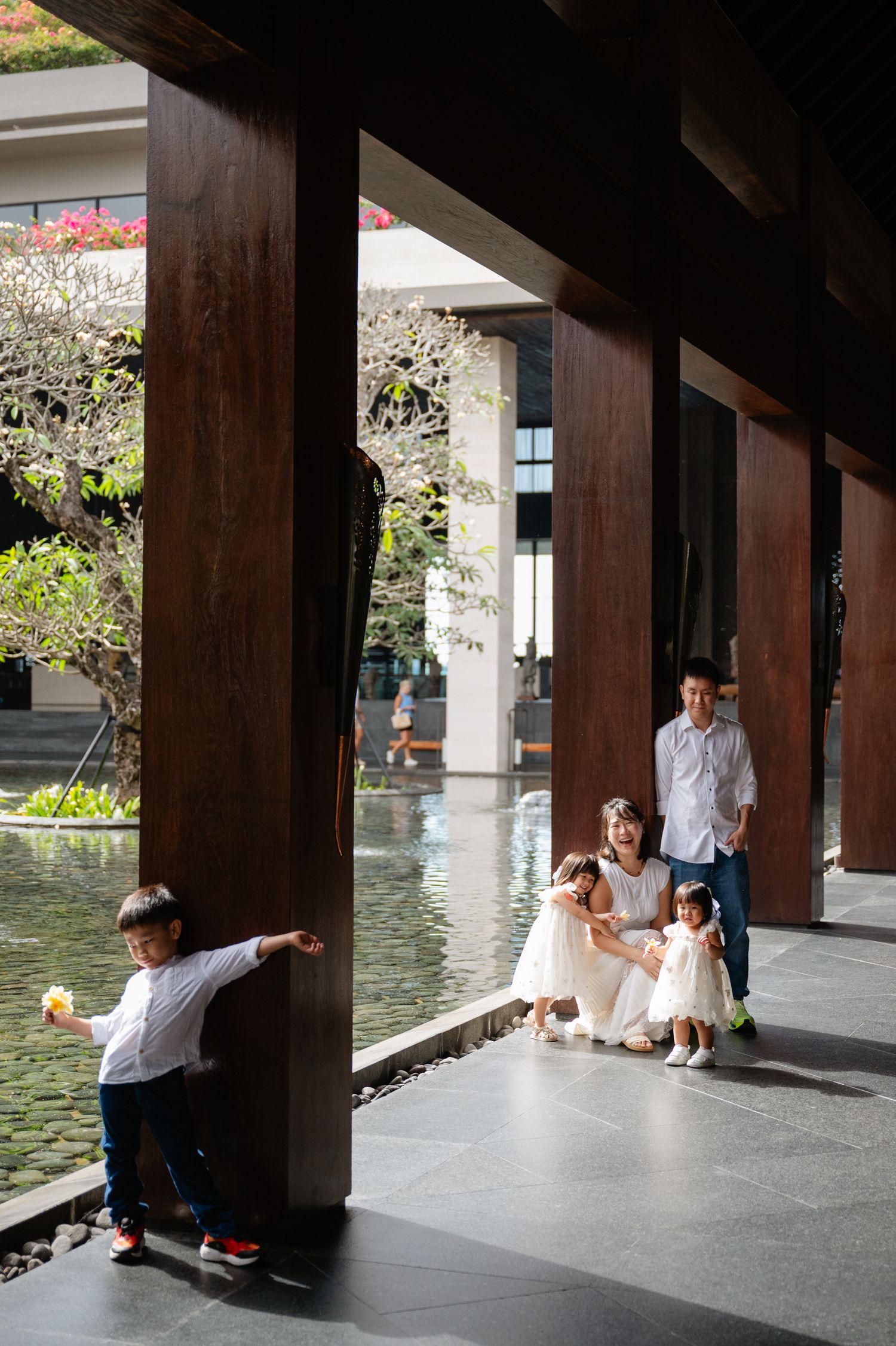 Family enjoying a serene courtyard with reflective pool, wooden columns, and lush greenery at a modern Asian resort.