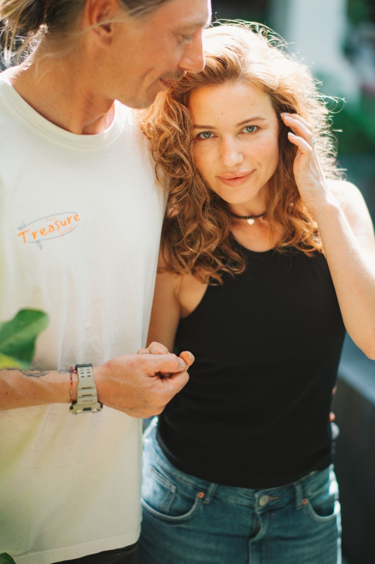 A young couple shares a tender moment outdoors, woman in black tank top leaning close to man in white Treasure tee.