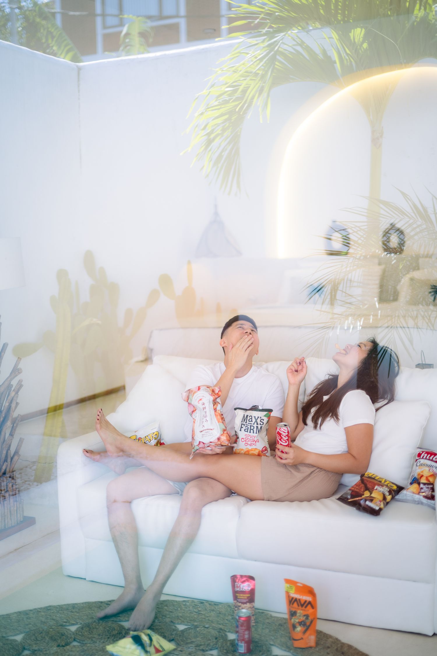 Couple relaxing on white sofa with snacks, laughing and enjoying leisure time indoors near tropical plants.