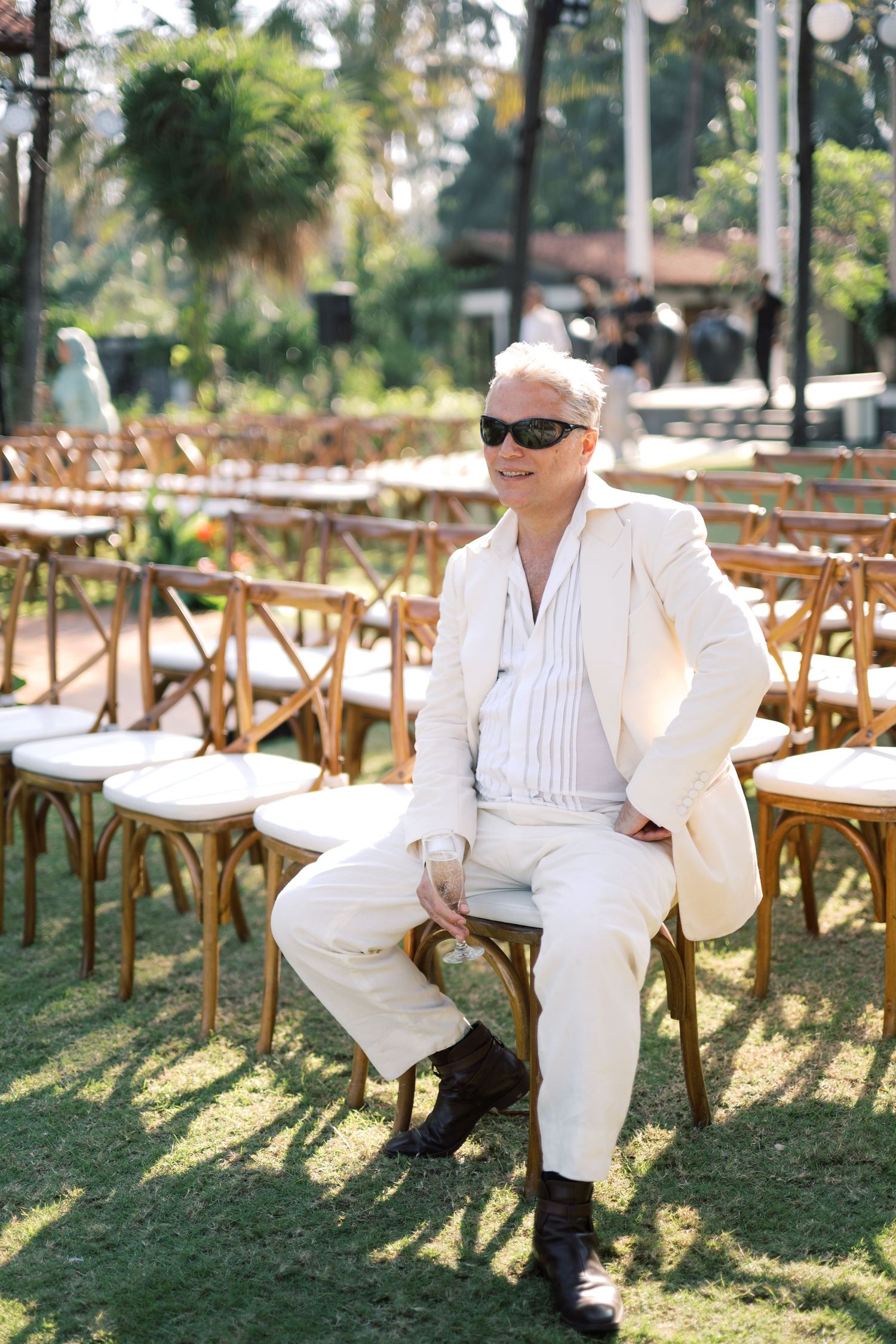 Stylish older man in white suit and sunglasses sitting on wooden chair at outdoor event venue with green lawn.
