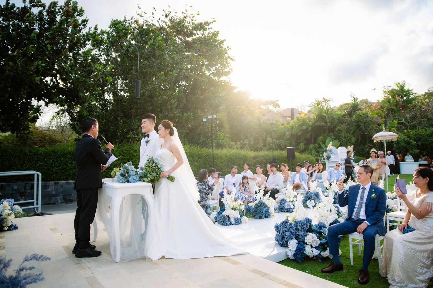 Bride and groom stand before an officiant at an outdoor garden wedding ceremony with guests seated among blue floral arrangements.