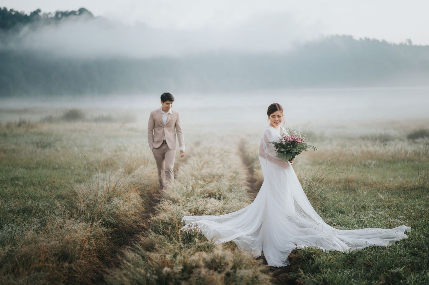 Bride in white gown and groom in beige suit pose on a misty meadow path surrounded by golden grass and fog.
