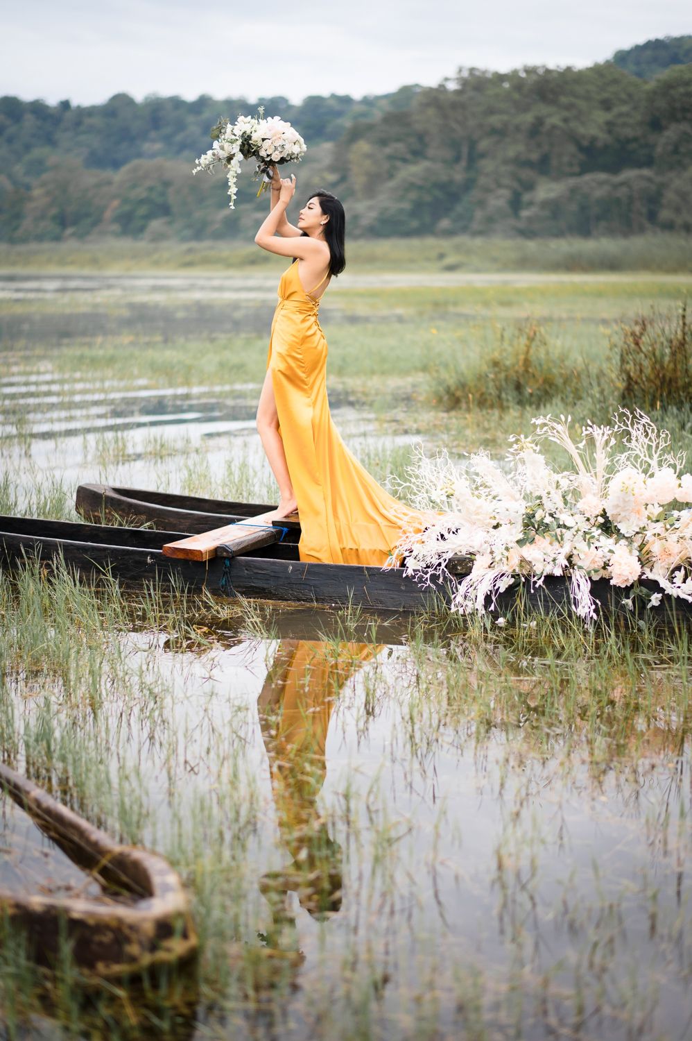 Woman in yellow gown standing on floral-decorated boat holding bouquet aloft over calm reflective waters.