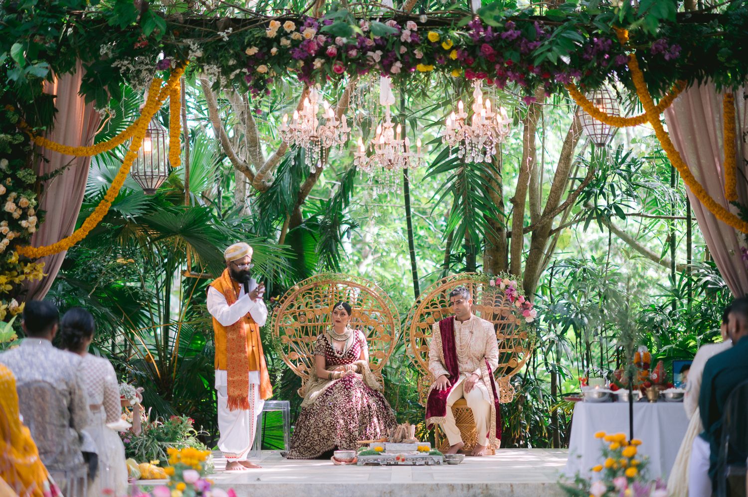 Elegant outdoor Indian wedding ceremony under floral arch with chandeliers and lush greenery.