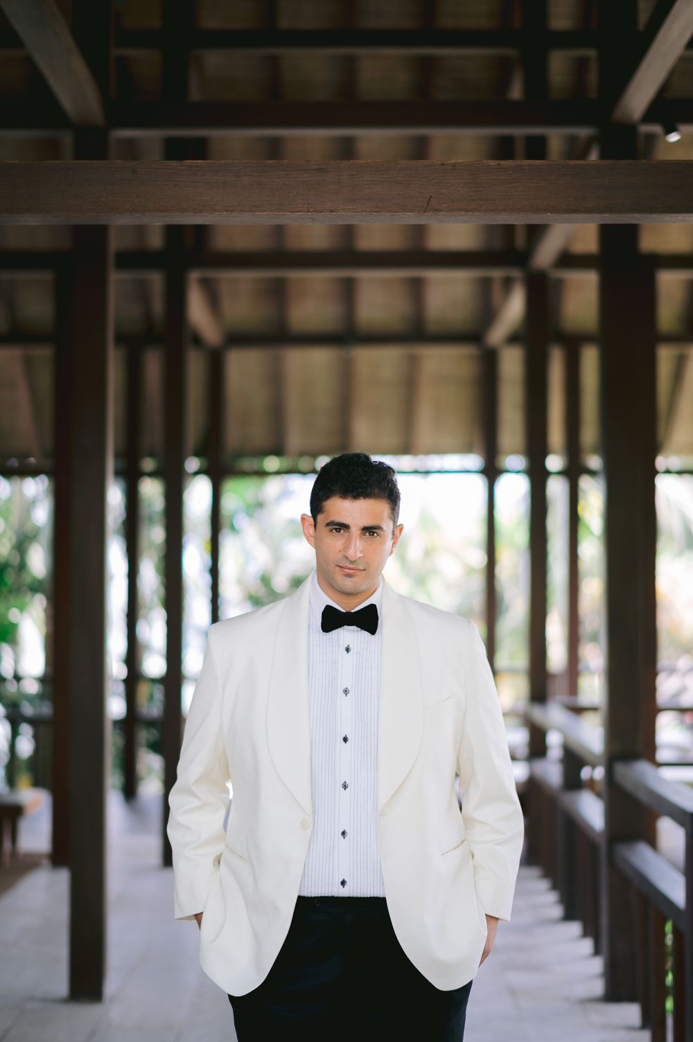 Groom in white tuxedo jacket and black bow tie stands in elegant open-air venue with wooden beam ceiling.