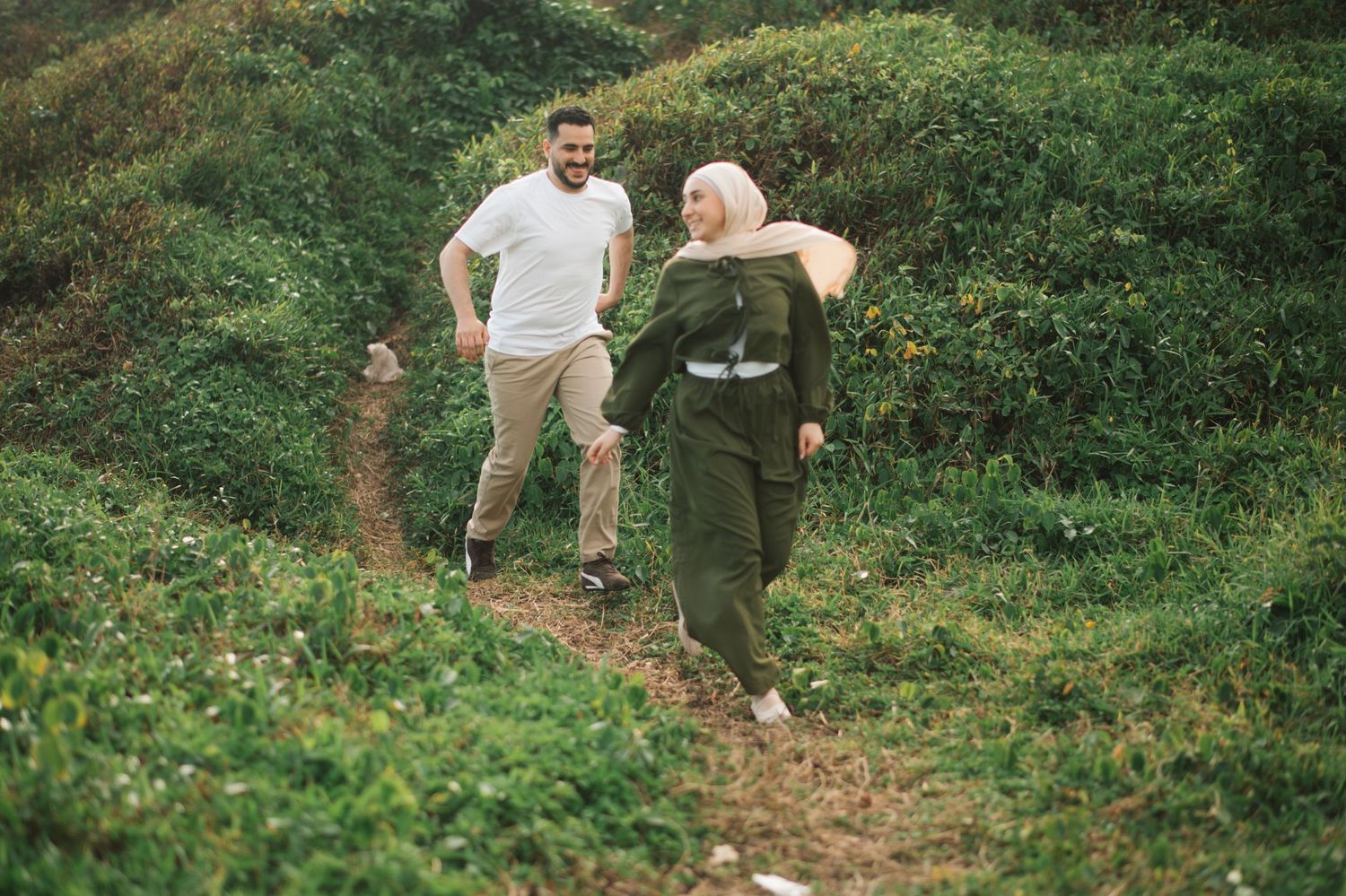 A couple joyfully hiking on a lush green trail, woman in hijab and green dress, man in white shirt laughing behind her.