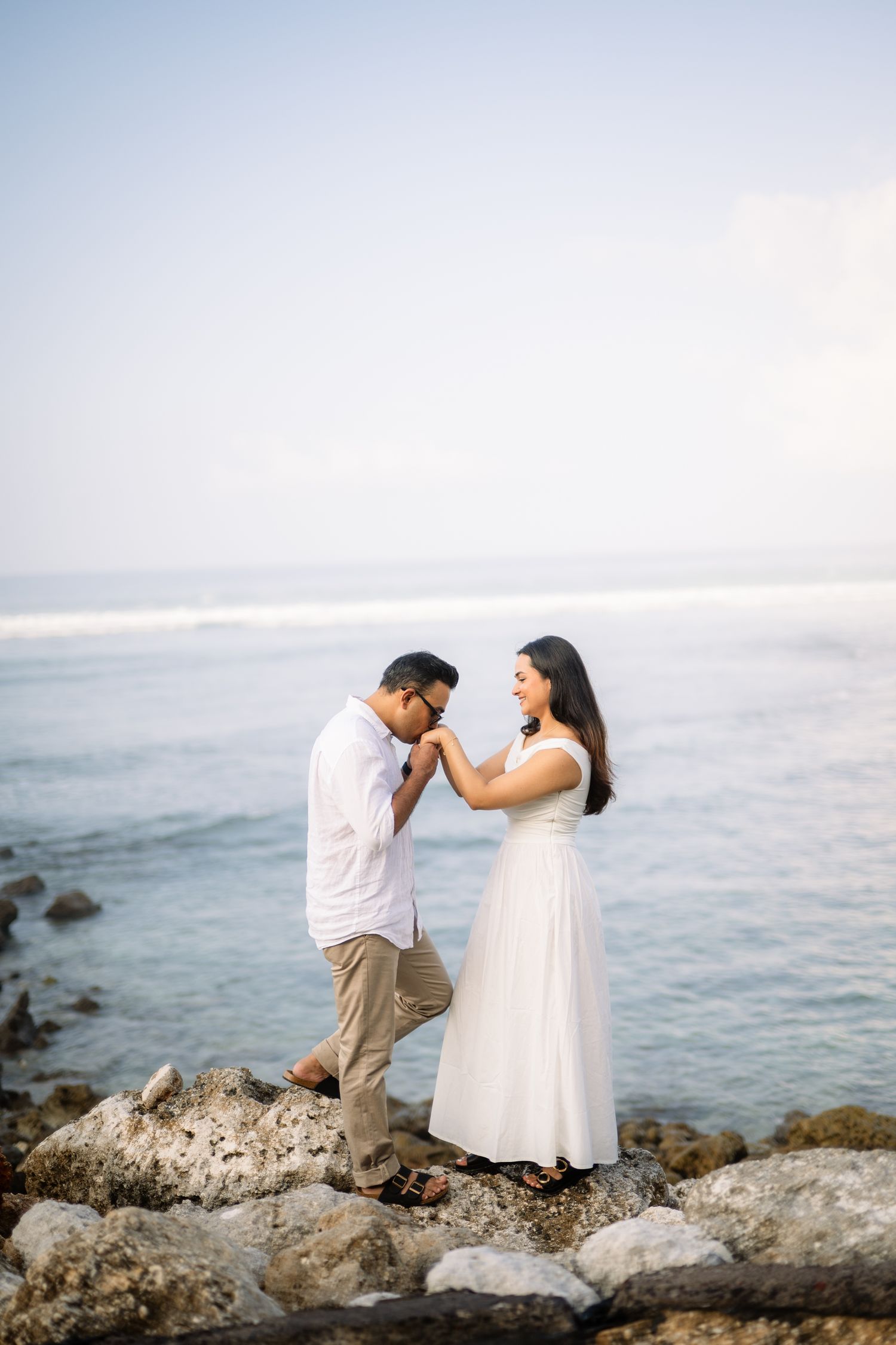 Romantic couple in white outfits standing on rocky shore, man kissing woman's hand with ocean backdrop.