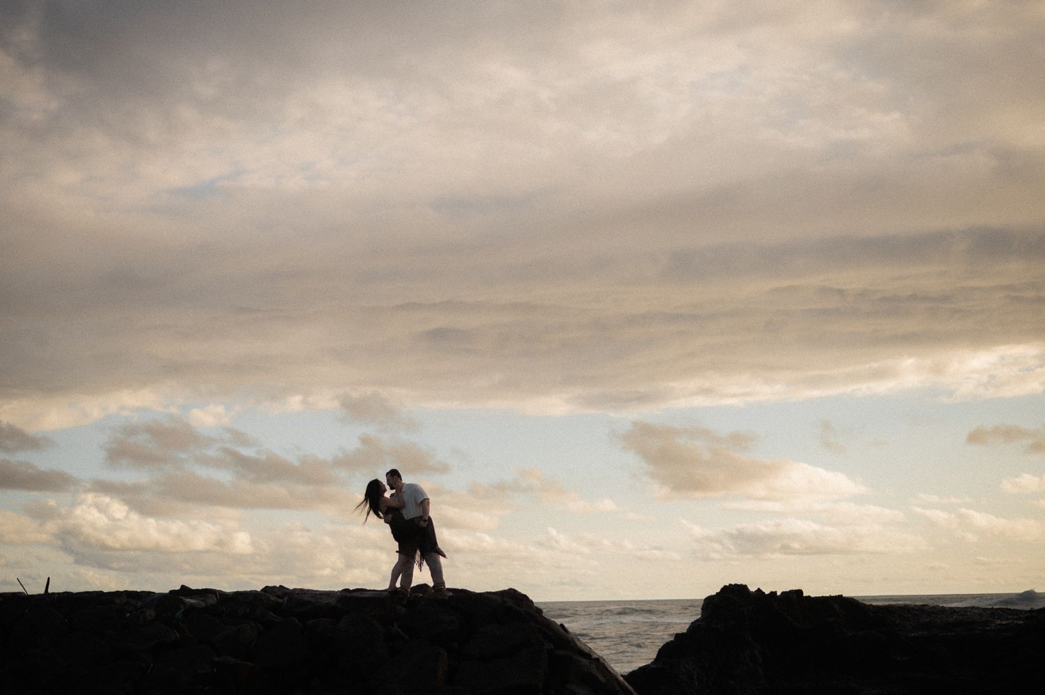 Silhouette of a couple embracing on rocky shoreline at sunset with dramatic cloudy sky backdrop.