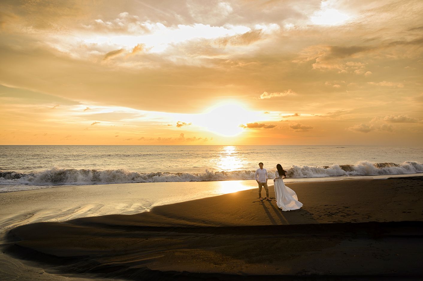 Couple in wedding attire walking on a sandy beach at golden sunset with ocean waves in the background.