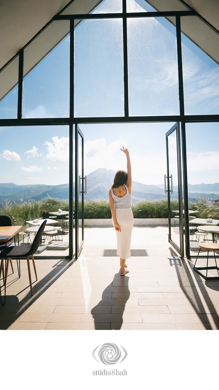 Woman in white dress raises arm toward panoramic mountain view through floor-to-ceiling glass doors in modern interior.