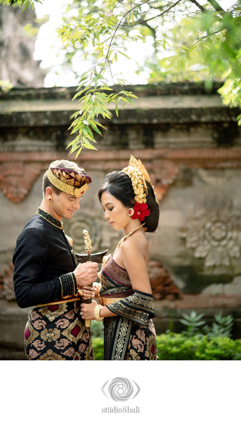 Couple in traditional Balinese wedding attire, man in black and gold, woman in ornate headdress, holding a ceremonial keris.