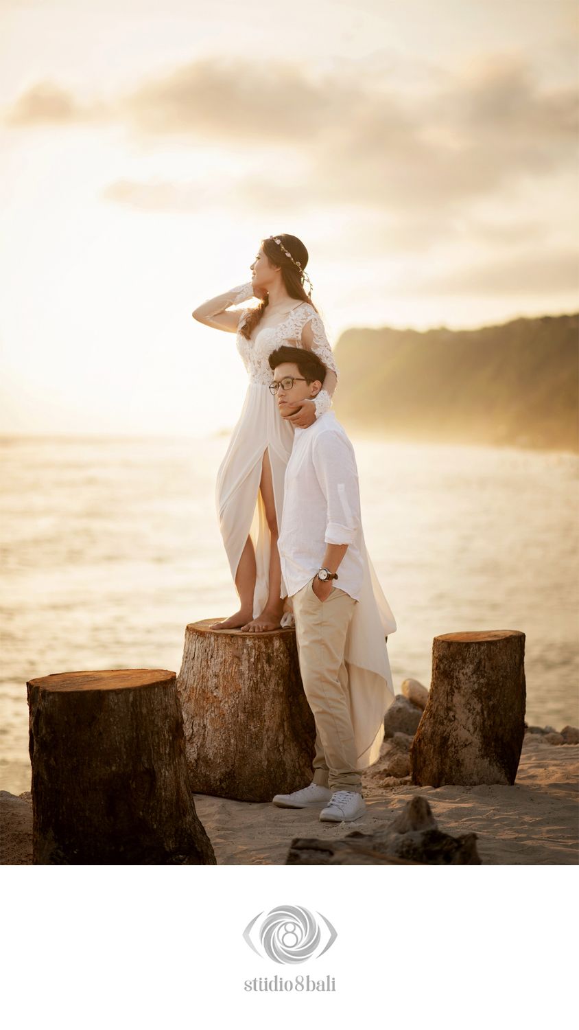 Couple in white outfits posing on tree stumps at a beach during golden sunset, romantic pre-wedding photo shoot.