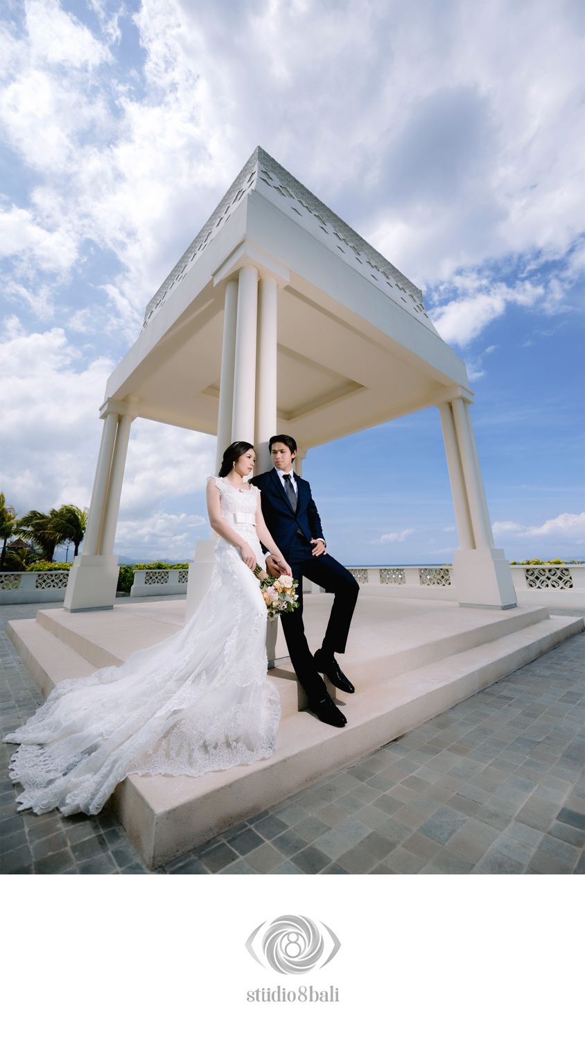 Bride in lace gown and groom in navy suit pose under elegant white pavilion with blue sky backdrop.
