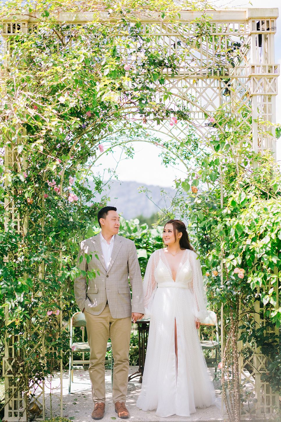 Couple holds hands under a floral garden arch, bride in white gown, groom in tan suit, lush greenery backdrop.