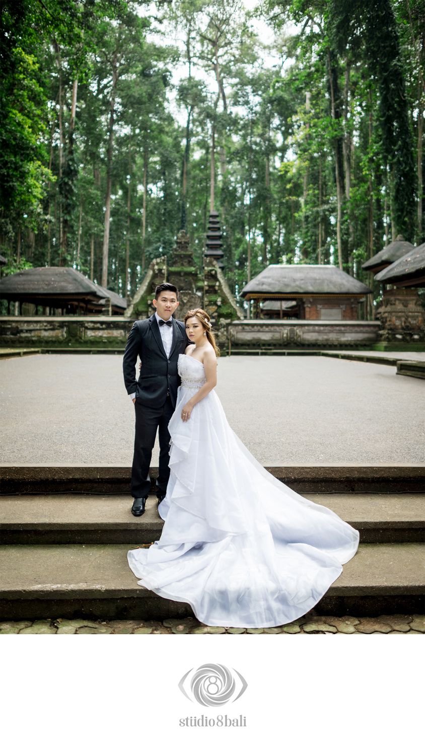 Couple in wedding attire pose on steps before Balinese temple surrounded by forest.