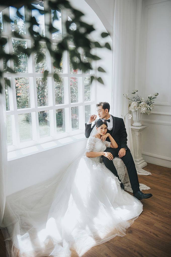 A couple in wedding attire poses by a bright window, the bride in a white gown and groom in a black suit.