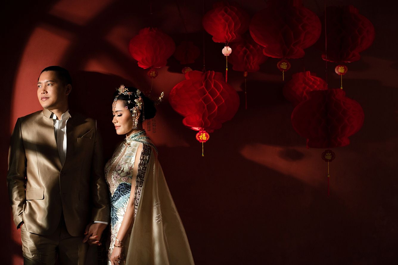 Couple in elegant traditional Chinese wedding attire poses before red lanterns in a dramatic, moody portrait.