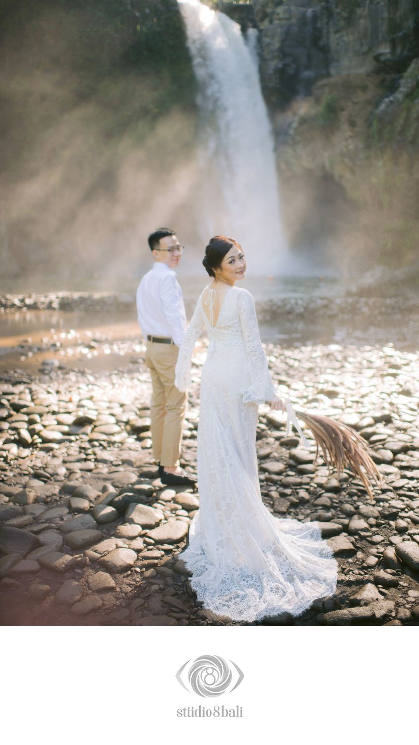 Couple poses before a majestic waterfall, bride in white lace gown holding dried flowers on rocky shore in Bali.