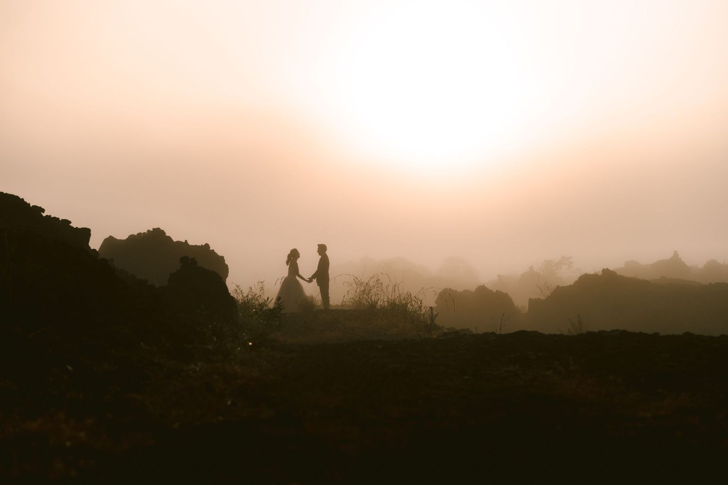 Silhouette of a couple holding hands at sunset in a misty rocky landscape, golden fog glowing behind them.