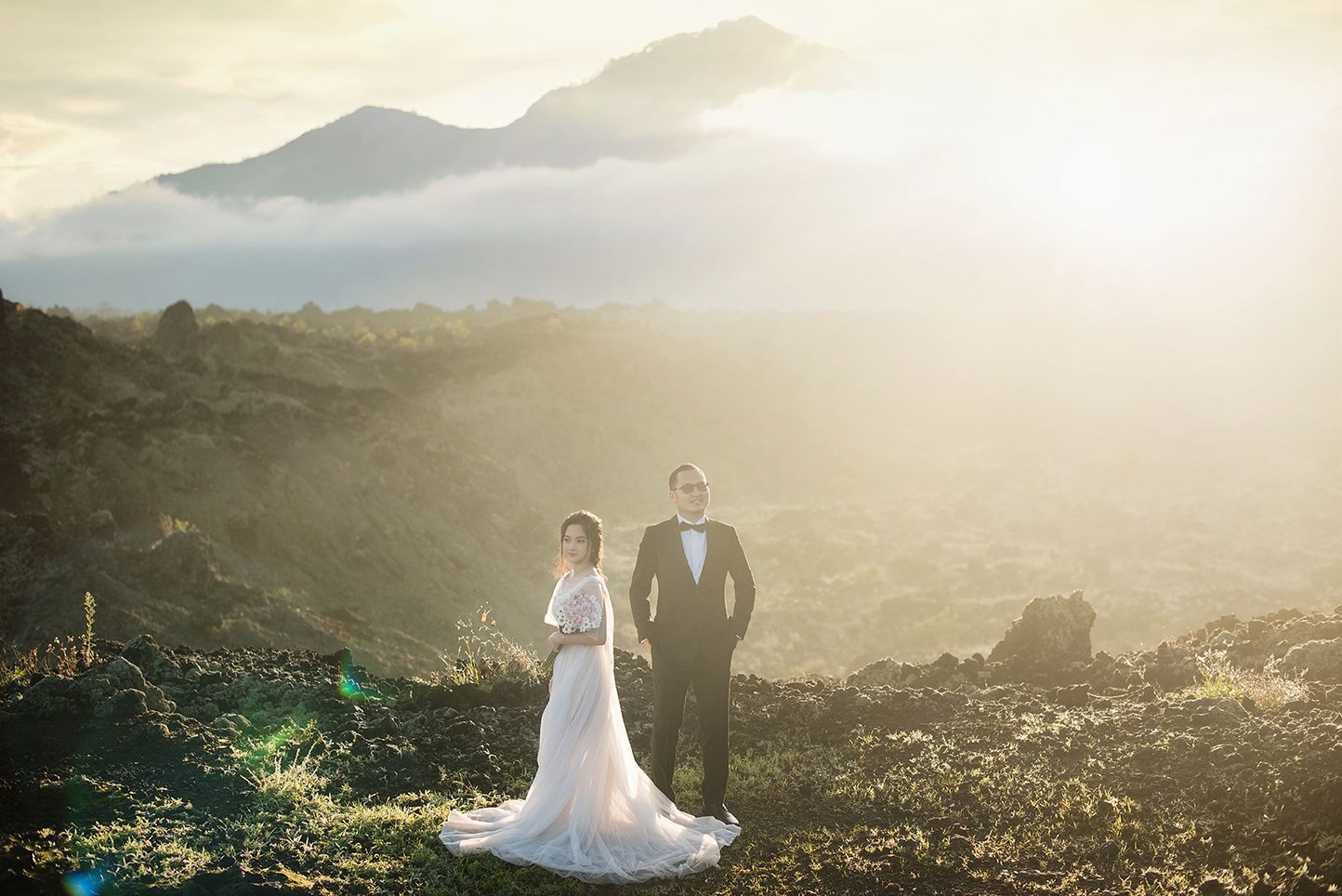 Bride and groom posing on volcanic mountain landscape at golden sunset with misty valley views.