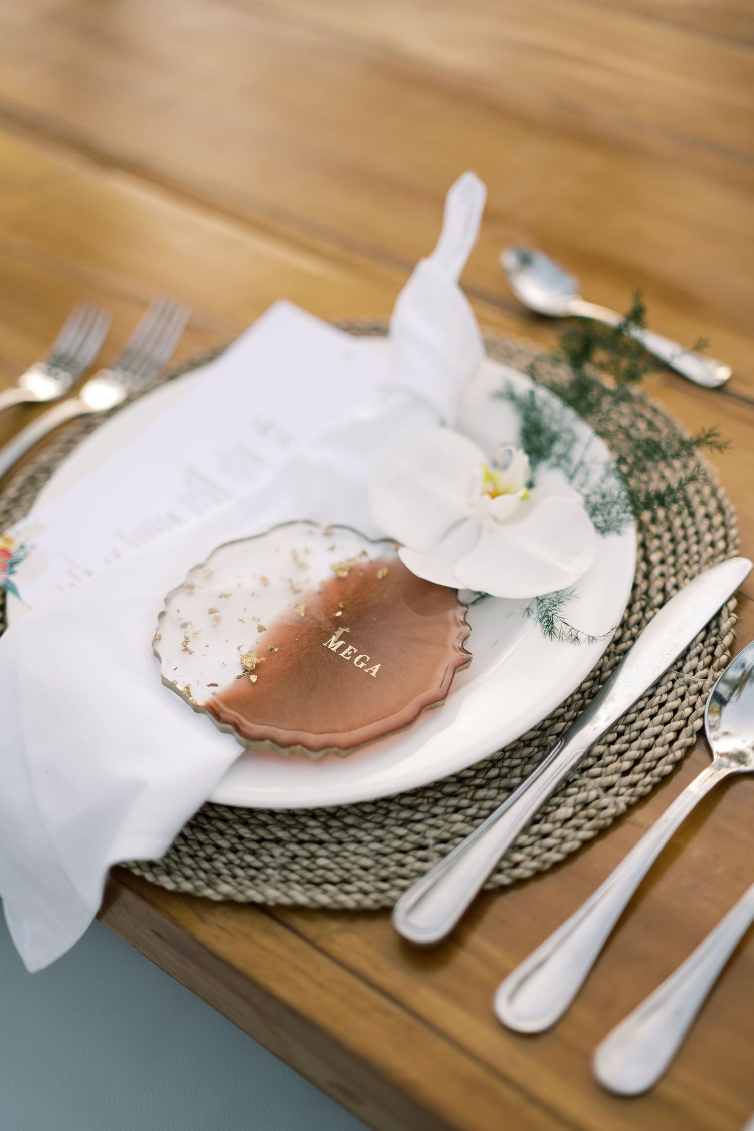 Elegant wedding place setting with woven charger, white orchid, copper name card, and silverware on wood table.