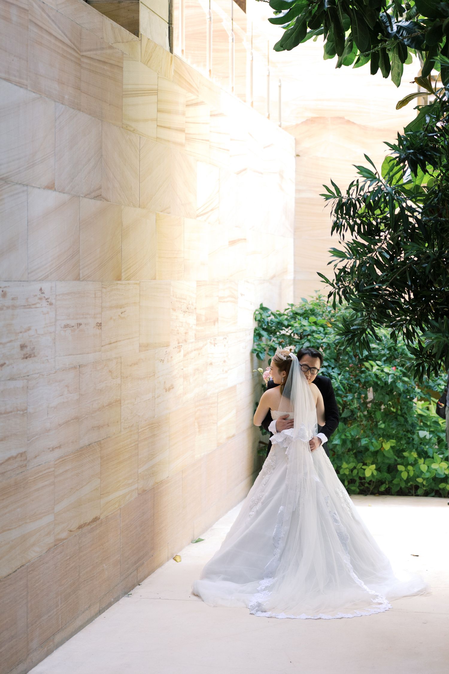Bride and groom sharing a romantic kiss against a sandstone wall, with lush green trees and a flowing white wedding gown.