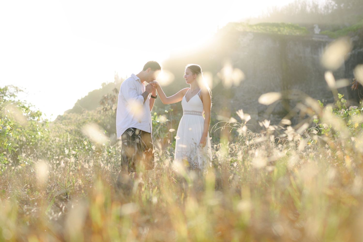 Couple holding hands in a sunlit meadow with wildflowers, sharing a romantic moment outdoors.