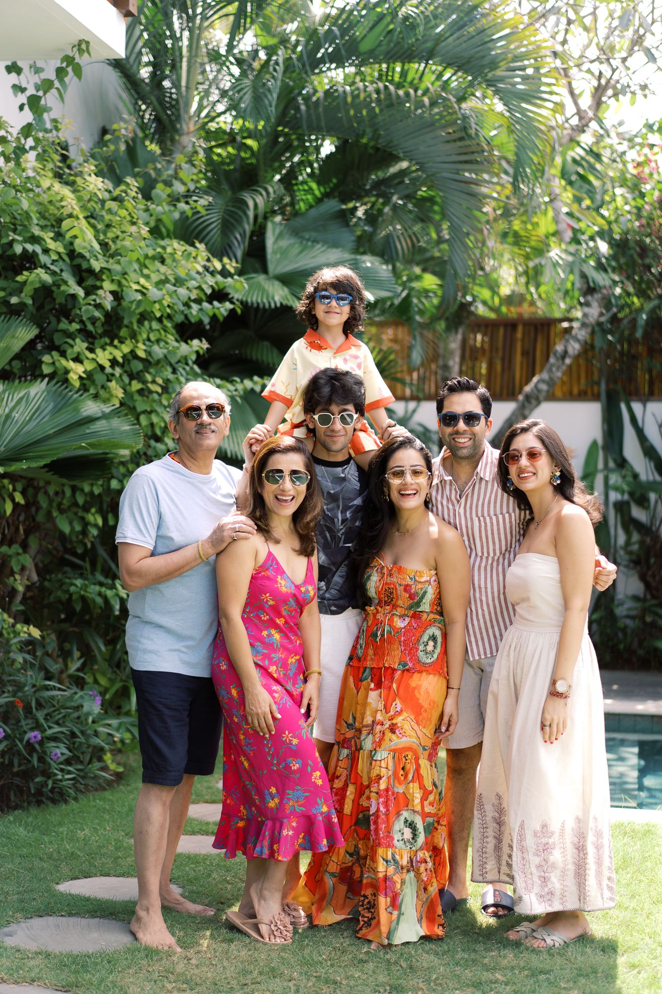 A joyful multigenerational family poses together outdoors in colorful summer outfits, with lush tropical greenery in the background.