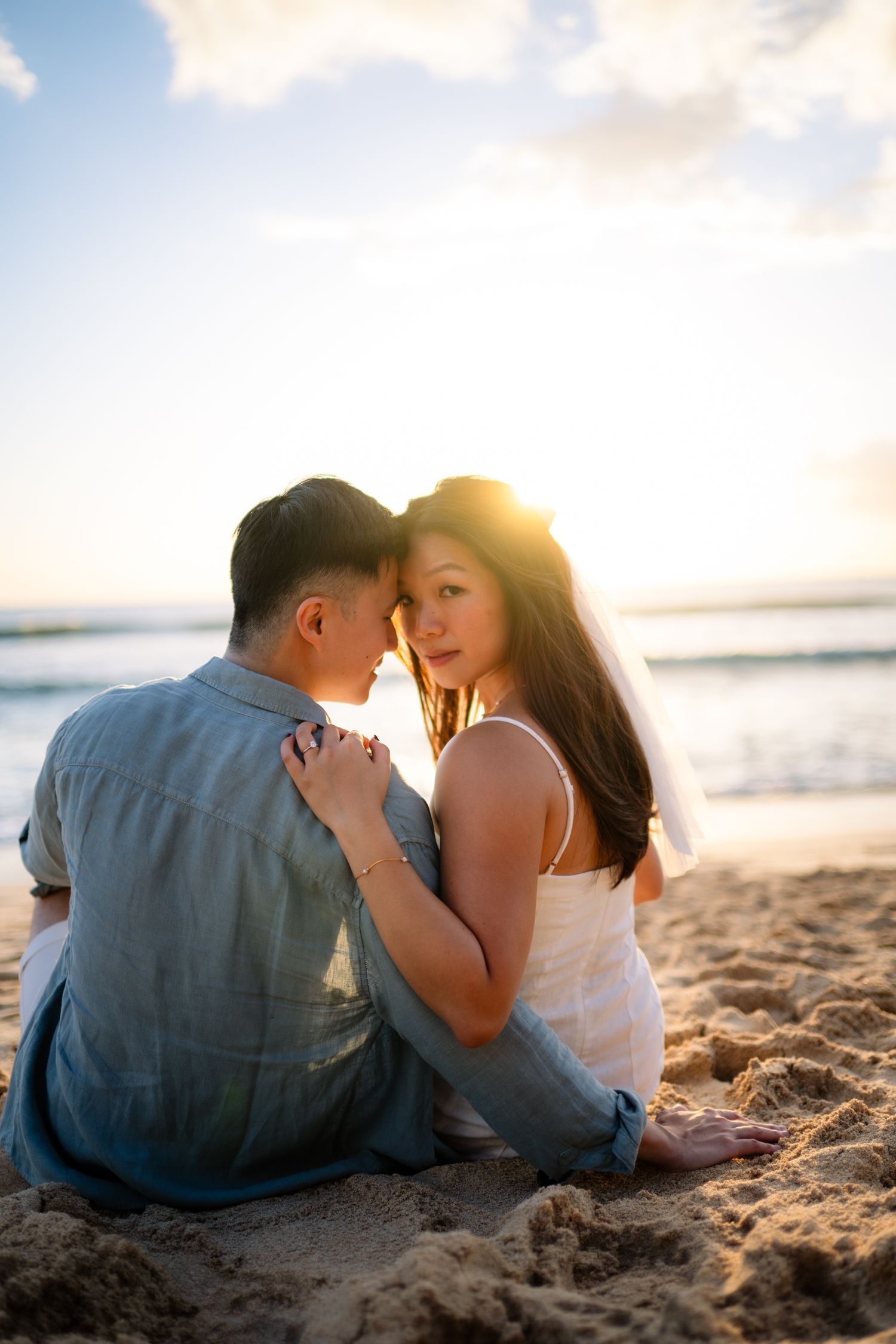 Bride and groom sitting on sandy beach at sunset, sharing a romantic moment with ocean waves in the background.