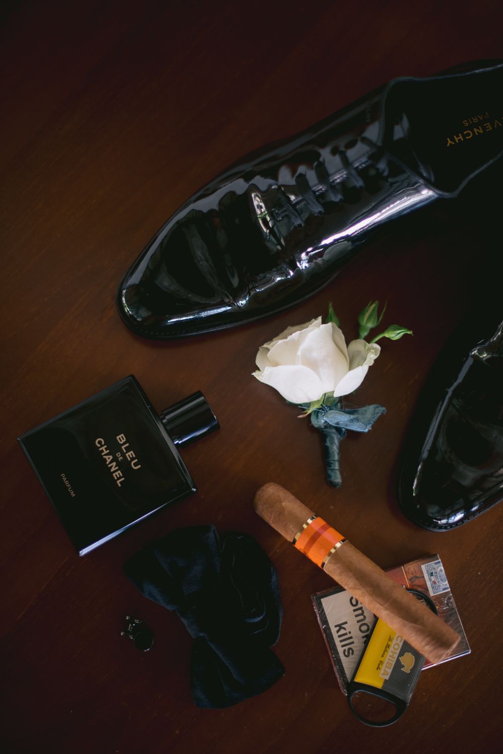 Groom's wedding accessories flat lay featuring Bleu de Chanel cologne, patent leather dress shoes, white rose boutonniere, and cigar.