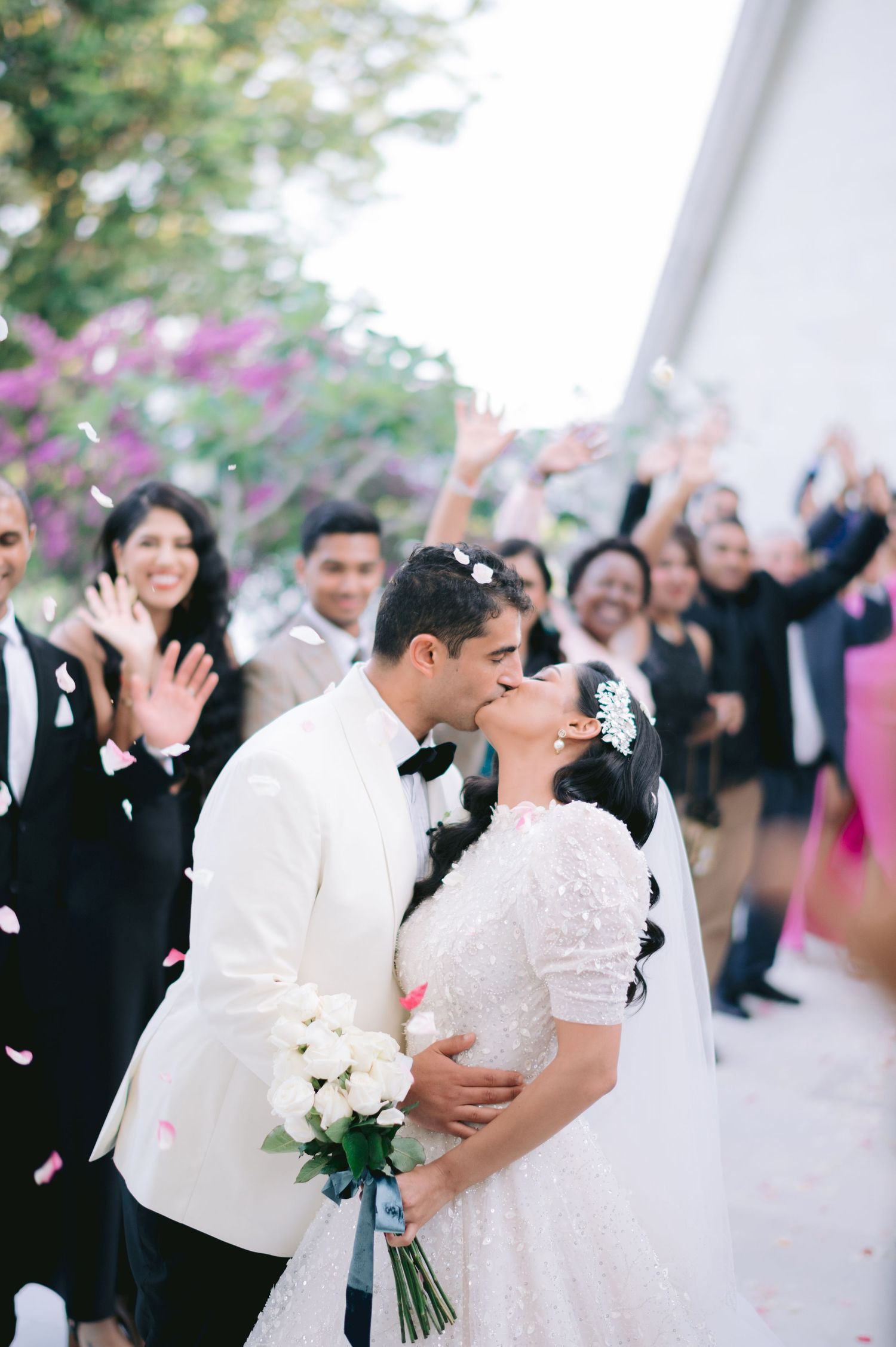 Bride and groom share a kiss as wedding guests celebrate with confetti and flower petals outdoors.