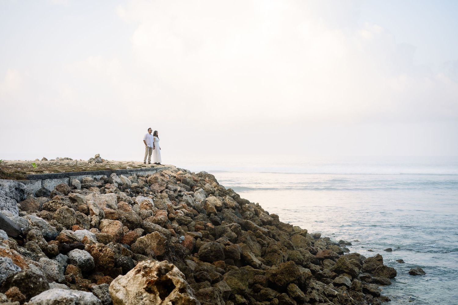 Couple standing on rocky coastal cliffs overlooking the ocean on a misty day.