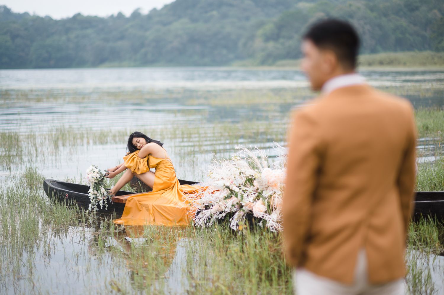 Woman in orange gown reclines in flower-adorned boat on misty lake while man in tan suit stands in foreground.