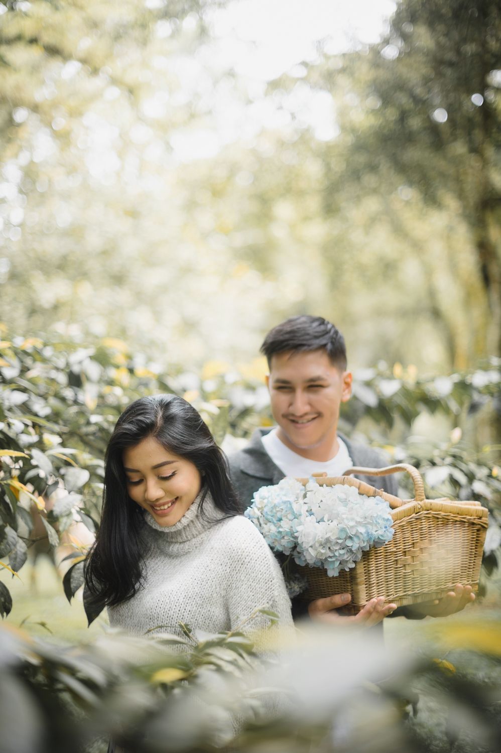Smiling Asian couple in a garden, man holding a wicker basket with blue hydrangeas, woman in a gray knit sweater.