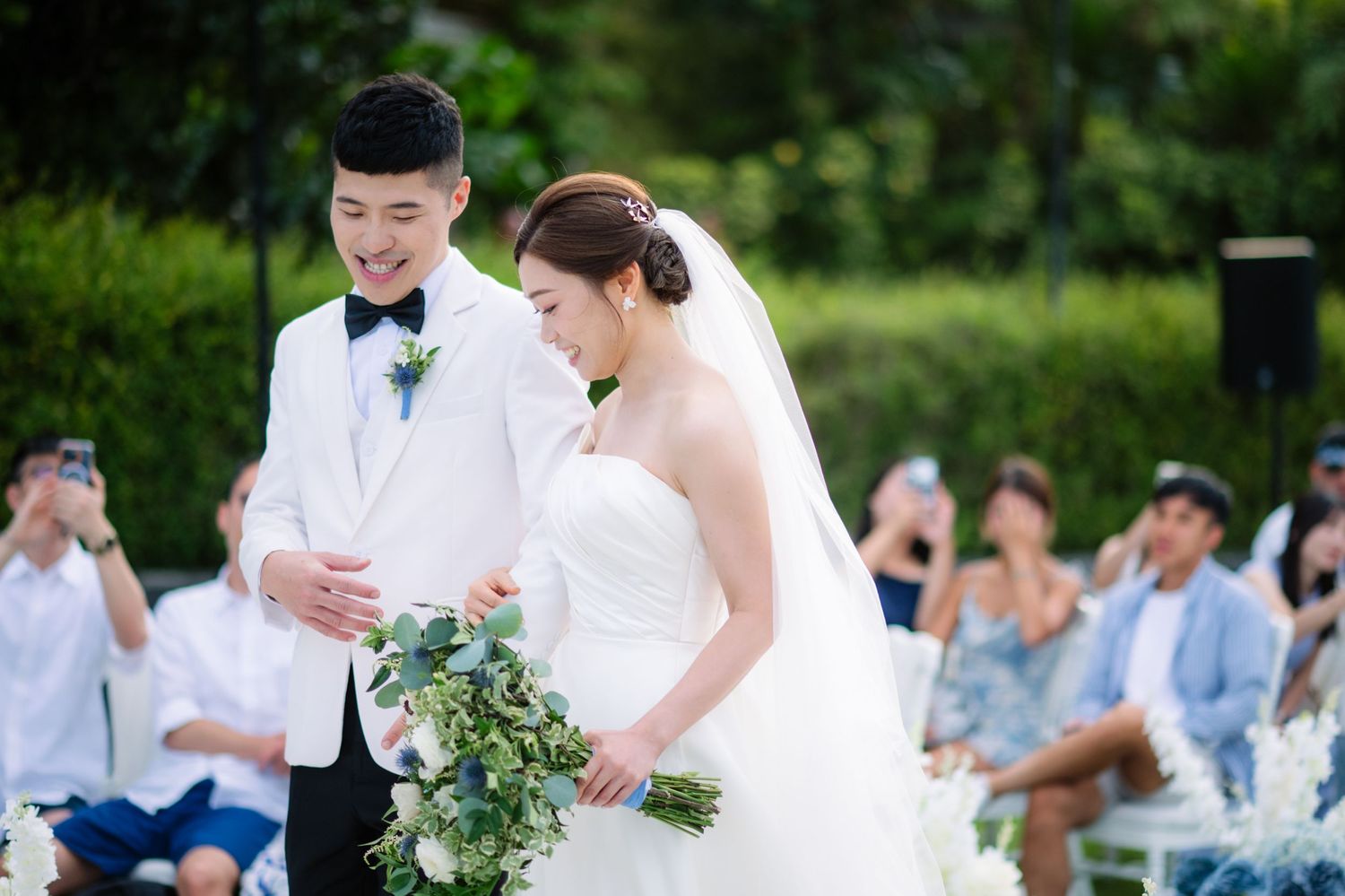 Bride in a strapless white gown and groom in a white suit hold a blue and white bouquet during an outdoor garden wedding ceremony.