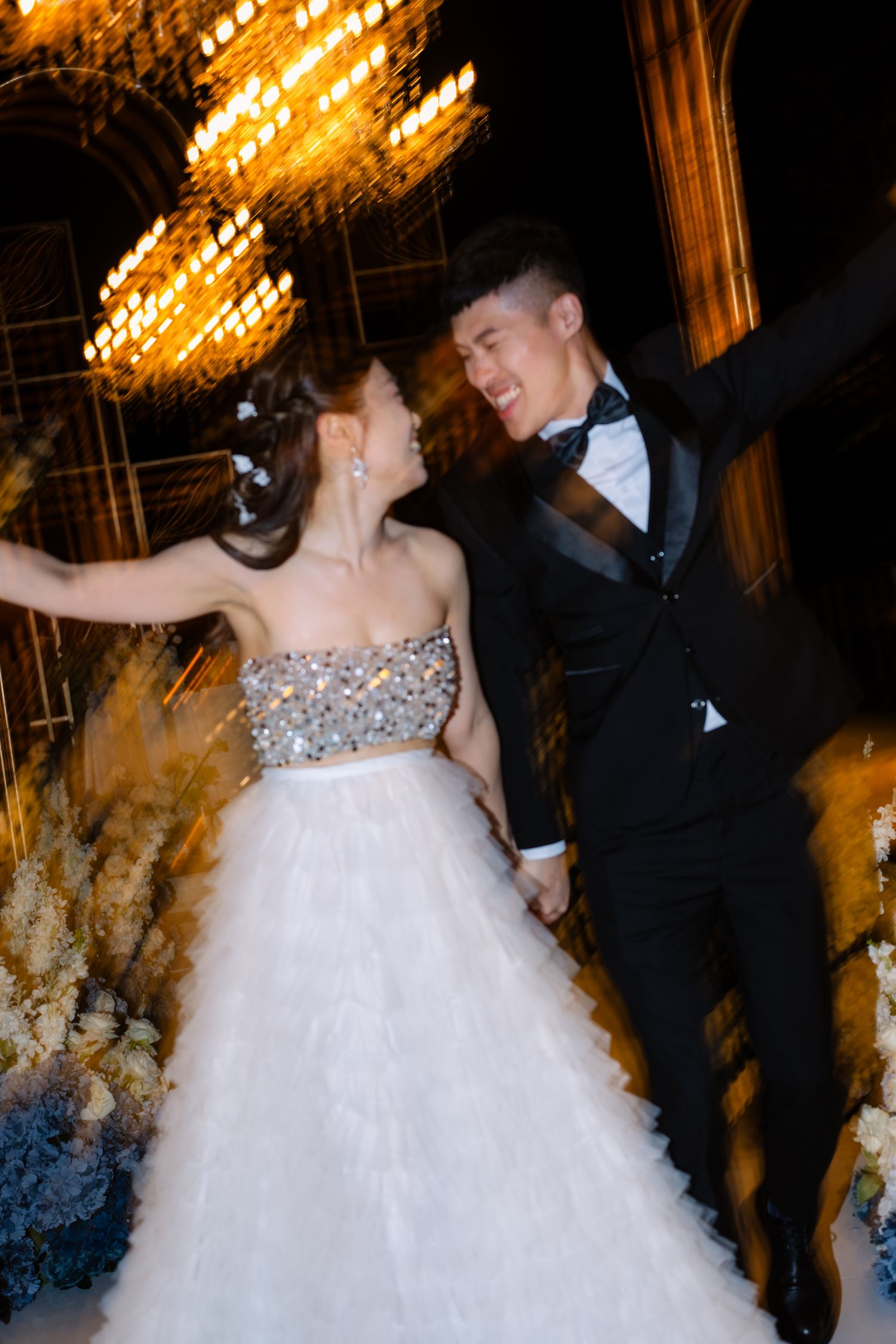Joyful bride in a sparkling strapless gown and groom in a black tuxedo dance under golden chandeliers at an elegant wedding reception.