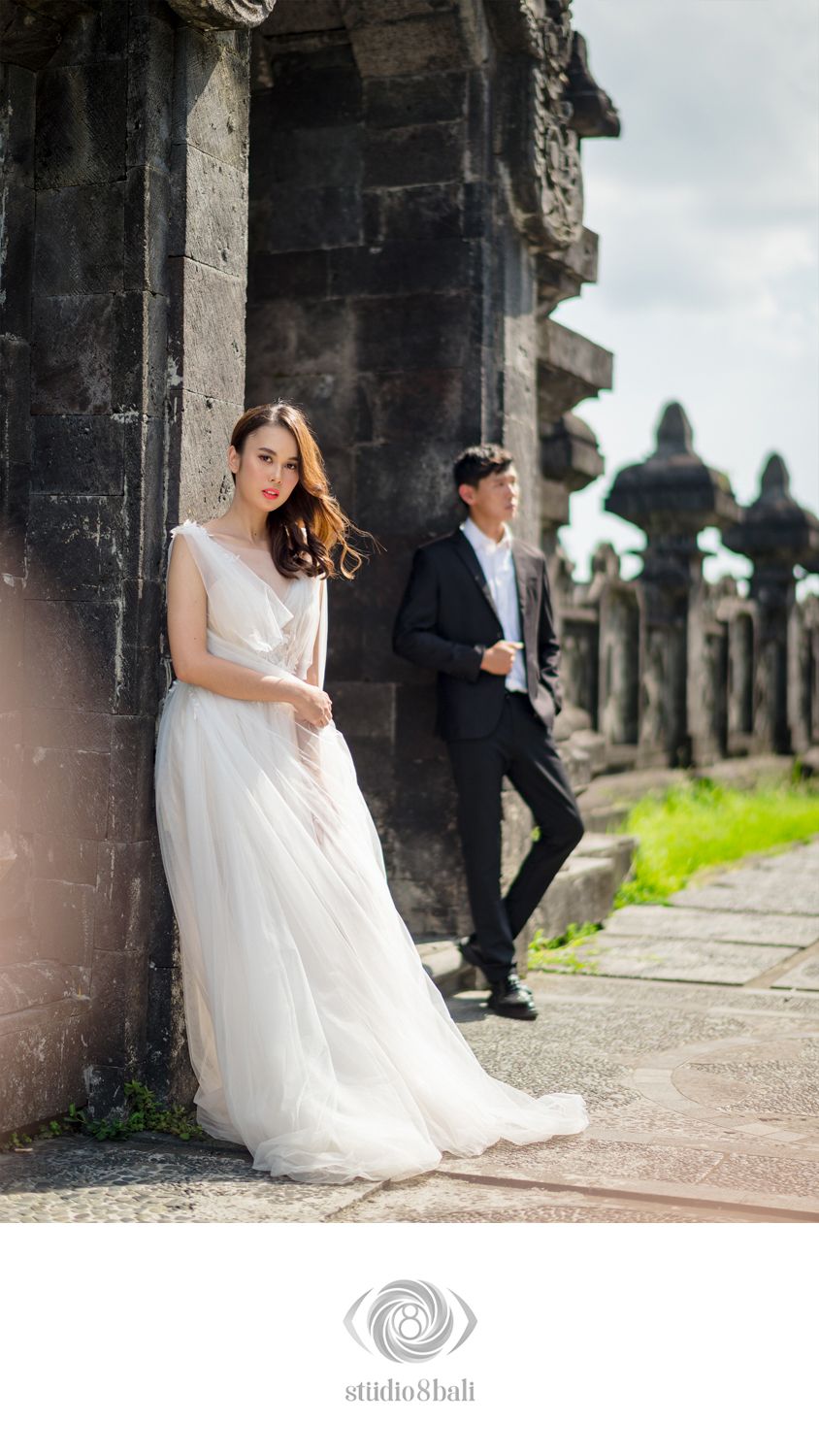 Couple in wedding attire posing at ancient stone temple, bride in white gown, groom in black suit, Bali.