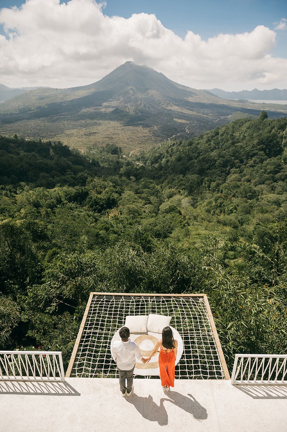 Couple standing on a rooftop net overlooking lush green jungle and volcanic mountain landscape in Bali.