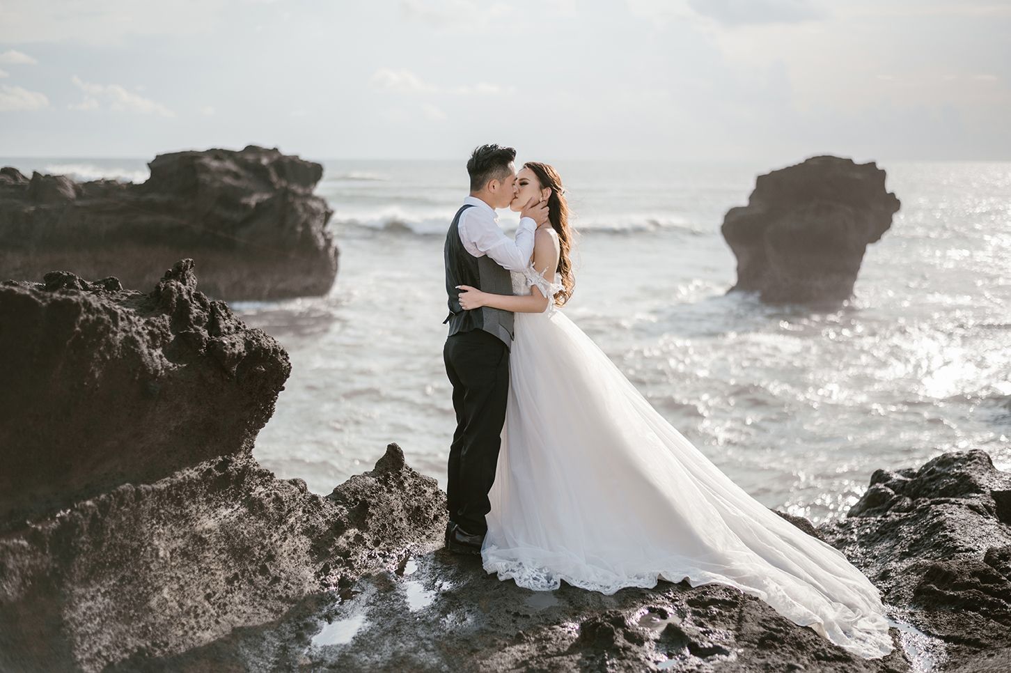 Couple in wedding attire embracing on rocky coastal cliffs with dramatic ocean waves and rock formations in background.