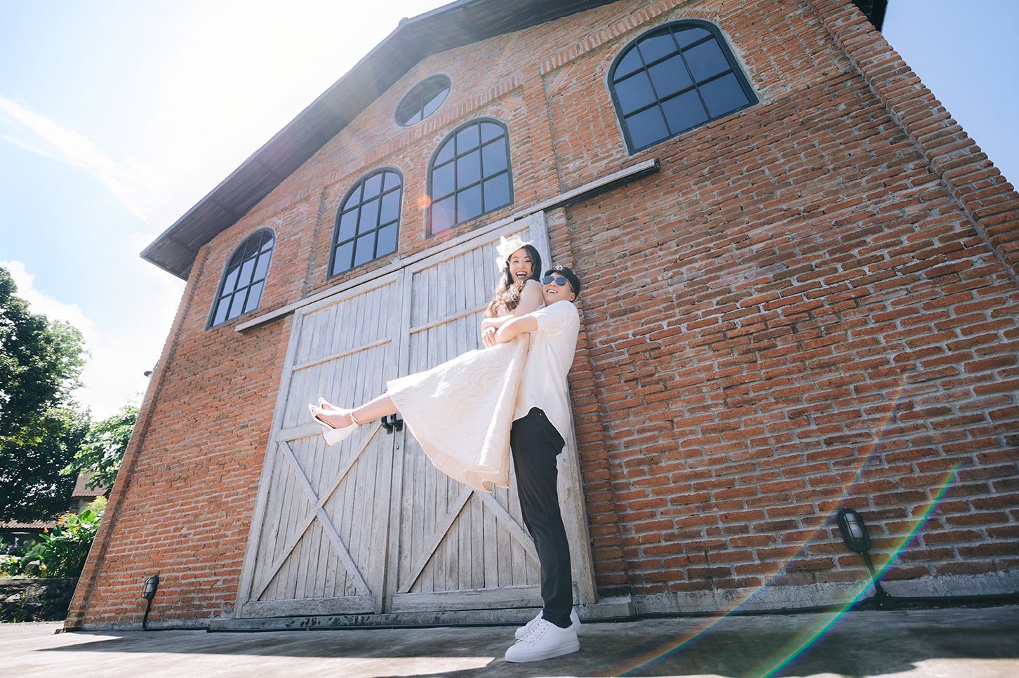Groom lifting bride in white dress outside rustic brick building with large wooden doors on sunny day.
