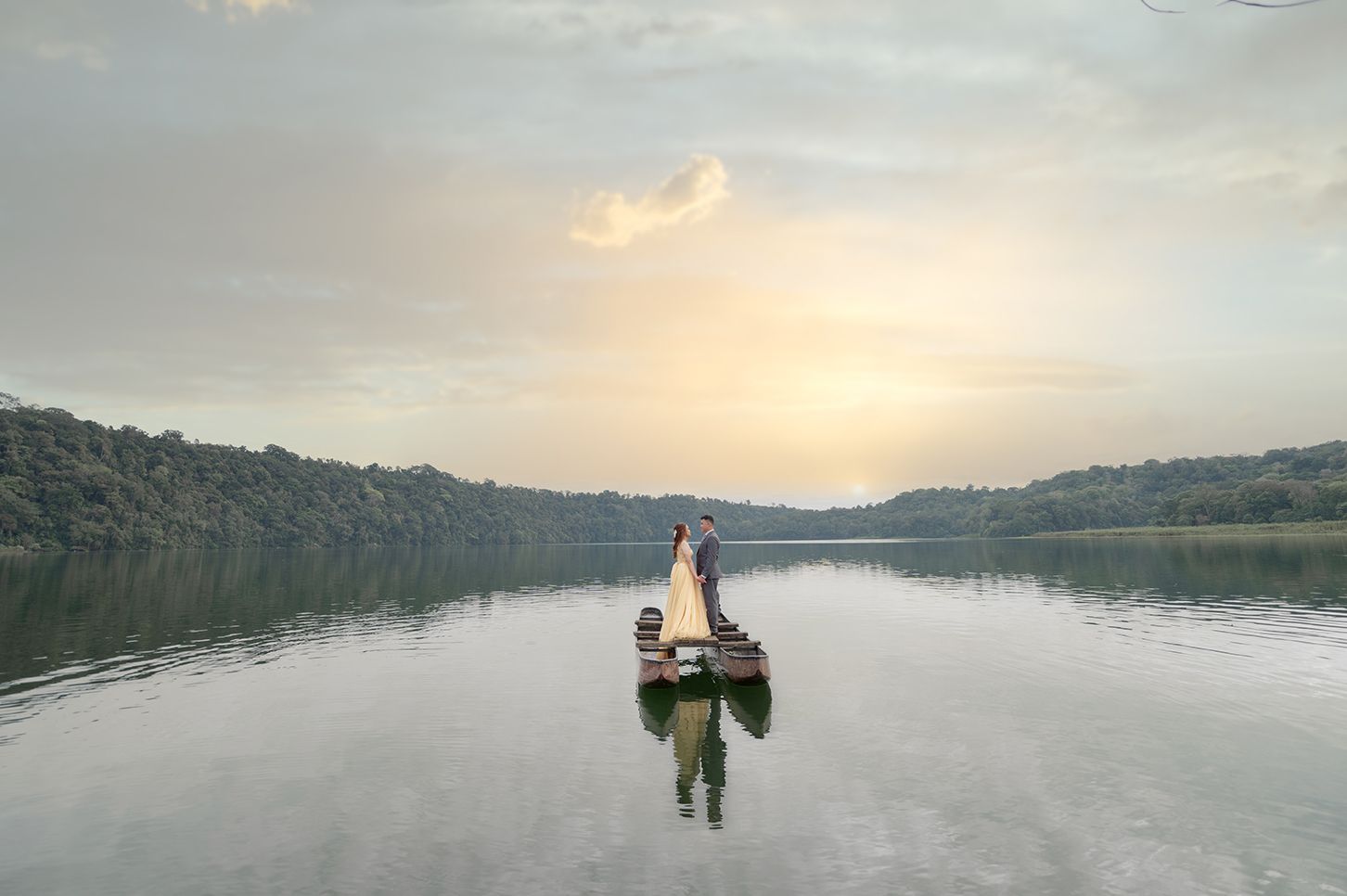 Couple standing on a wooden boat on a calm lake at sunset with lush green trees and golden sky reflected in water.