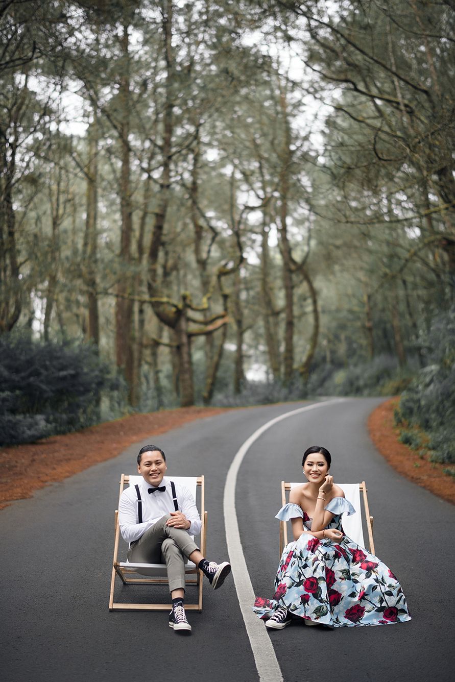 Couple in formal attire sitting in deck chairs on a tree-lined road for a creative pre-wedding photo shoot.