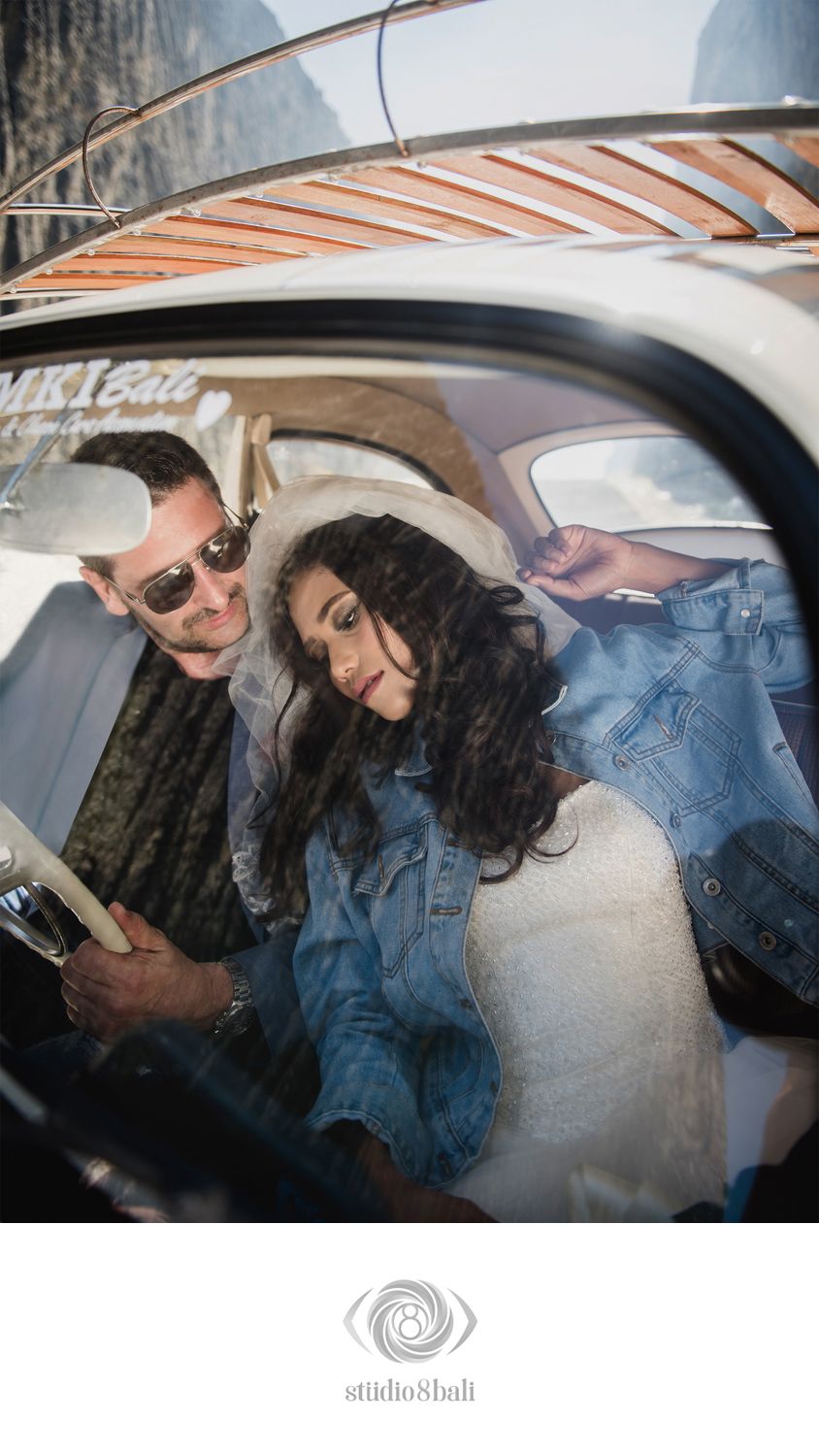 Couple sitting inside a vintage car, man wearing sunglasses, woman in denim jacket, photographed by Studio 8 Bali.